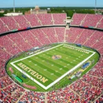 Aerial view of packed college football stadium during afternoon game with thousands of fans in colorful team apparel, sunny weather, vibrant atmosphere