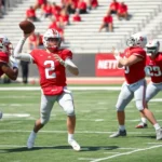 Professional college football players in scarlet and gray uniforms executing a precise passing play during spring practice, quarterback releasing the ball with perfect form while receivers run crisp routes in a sunlit stadium